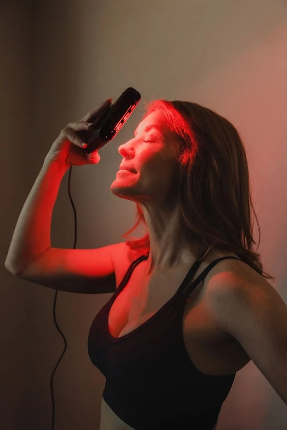Woman using a black handheld device on her forehead, emitting red light, in a dimly lit room.