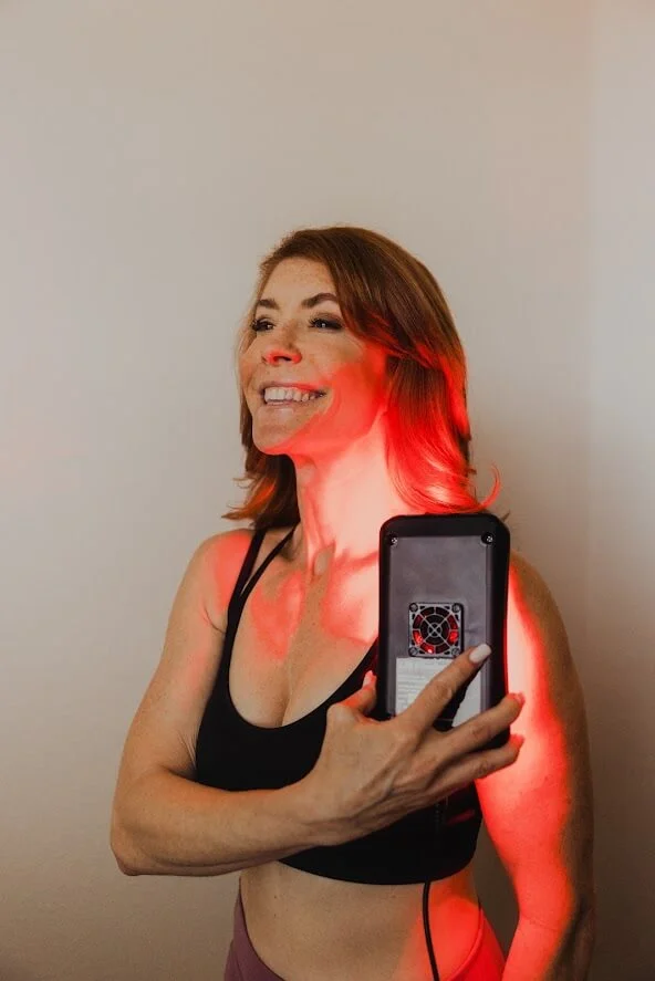 A smiling woman uses a handheld red light therapy device on her upper arm, standing against a neutral wall.