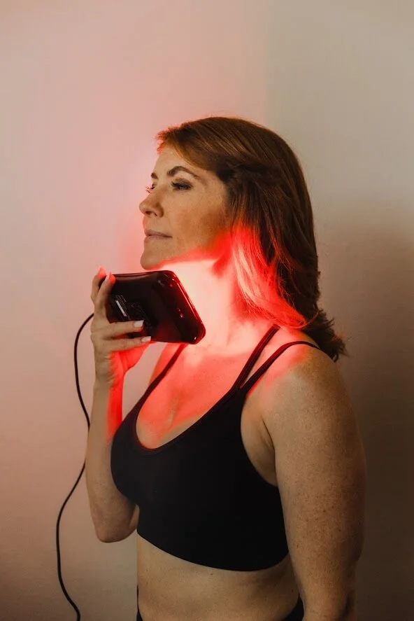 A woman uses a handheld device emitting red light on her neck, aimed at red light therapy, wearing a black tank top.