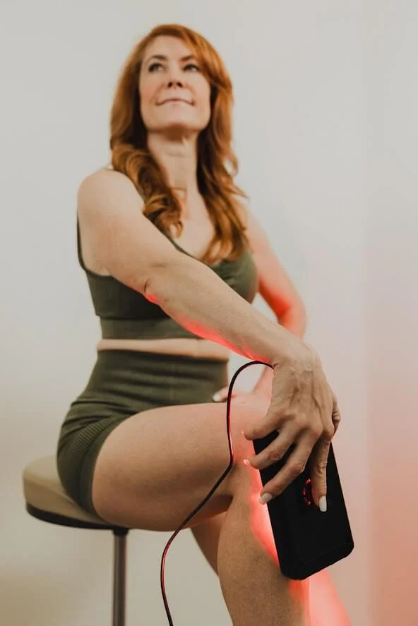 A woman in athletic wear uses a device on her leg, emitting red light, while sitting on a stool.