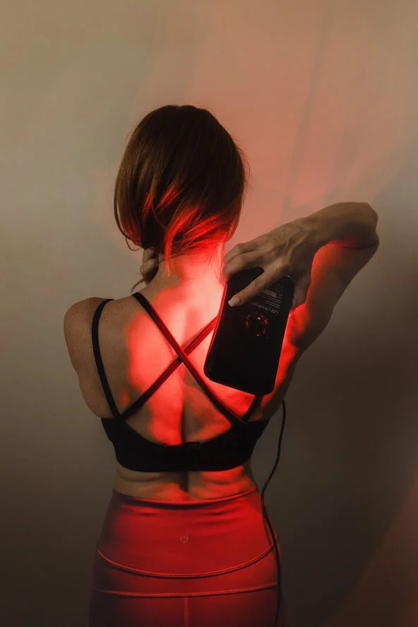 A person in a black sports bra uses a handheld device on their back, illuminated by red light.