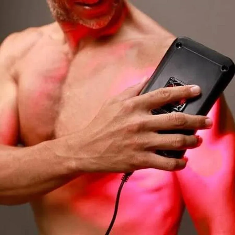 A shirtless man uses a handheld red light therapy device on his chest, which has a warm glow from the device.