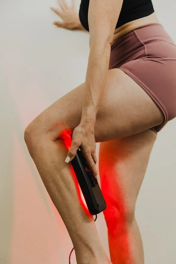 A person using a handheld red light therapy device on their thigh, set against a neutral wall.