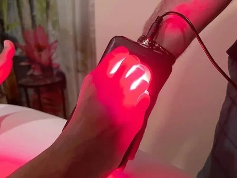 A hand holds a device emitting red light, positioned above another hand, possibly for therapeutic use.
