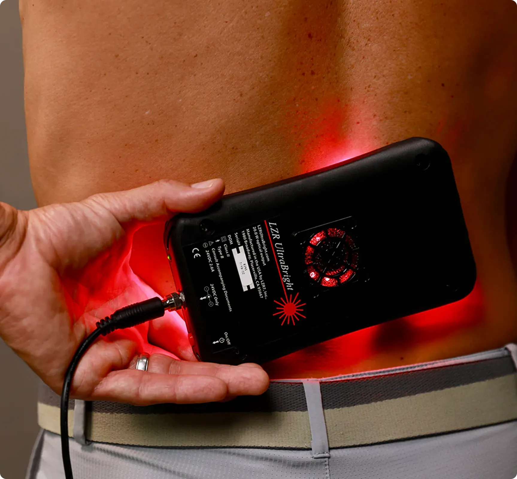 A person holds a handheld device with red lights against their lower back, likely for therapeutic purposes.