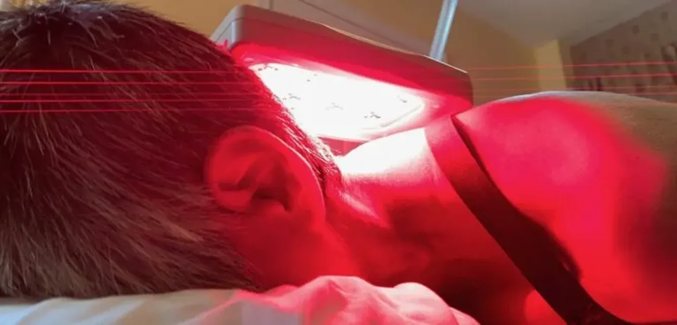 A person lies face down on a bed, receiving red light therapy from a device positioned above them.