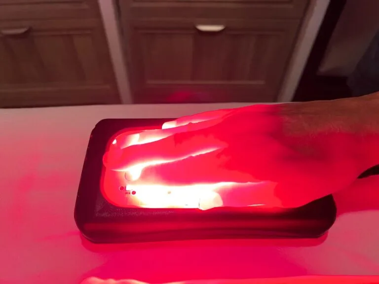 A hand resting on a device emitting red light, for therapeutic treatment, against a blurred background.