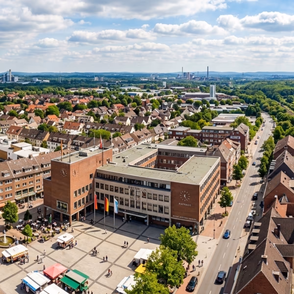 Luftaufnahme eines belebten Rathausplatzes mit einem großen Gebäude, umgeben von Wohnhäusern und grünen Bäumen unter einem bewölkten Himmel.