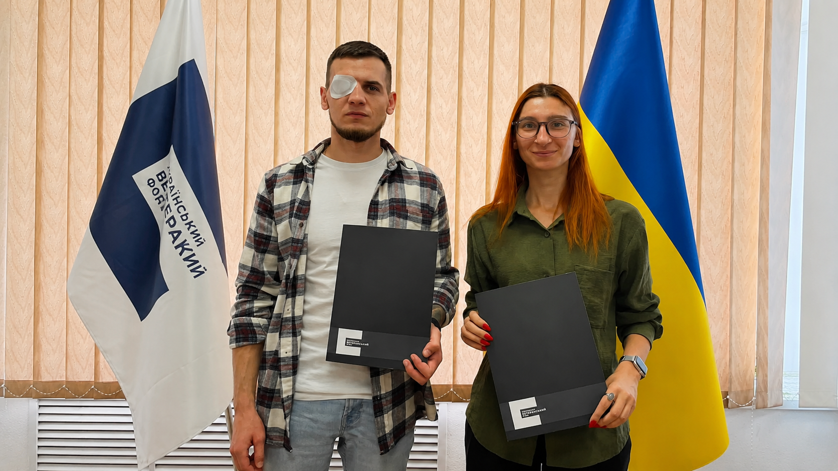 Представники КІБЕРАКАДЕМІЇ SHERIFF та Українського ветеранського фонду після підписання меморандуму про співпрацю, тримають папки з документами на тлі прапорів України та Українського ветеранського фонду.