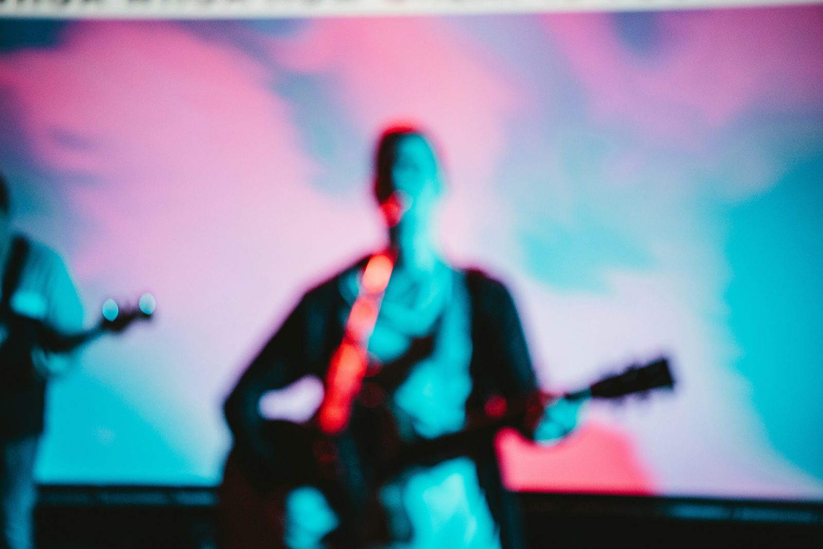 Blurred musician playing guitar on stage under pink and blue lights.