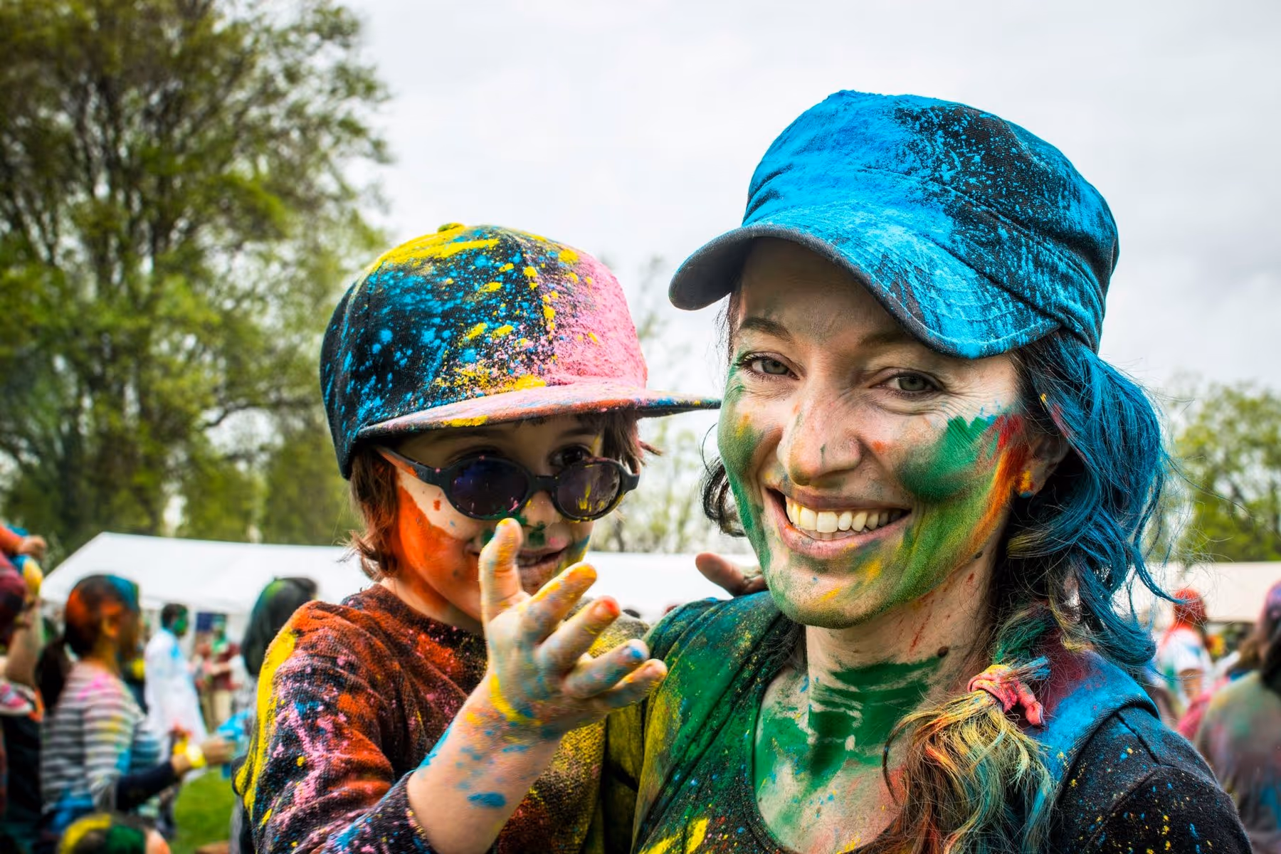 A woman and child, covered in vibrant colored powders, smile joyfully at a festival. They wear colorful caps and the background shows people and trees.