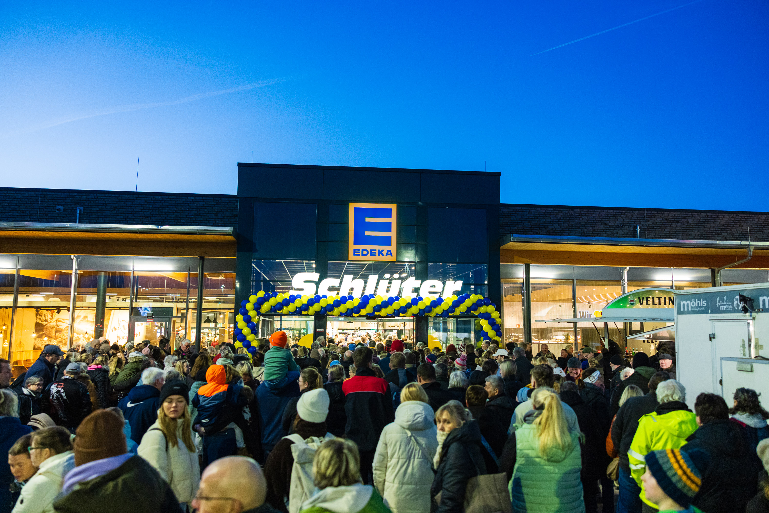Große Menschenmenge vor dem beleuchteten Eingang eines EDEKA-Schlüter Supermarkts bei Abenddämmerung.