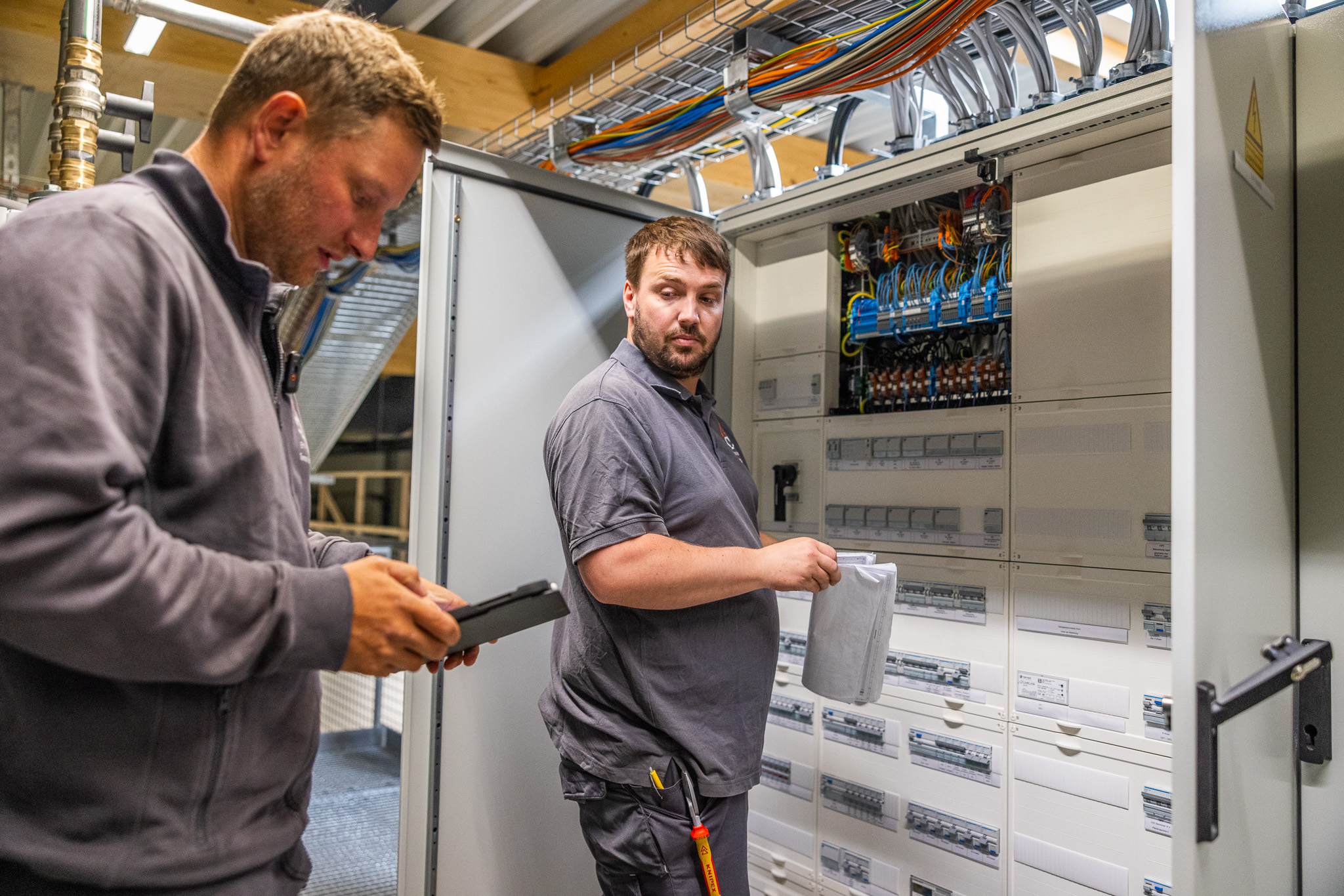 Zwei Techniker prüfen Kabel und Pläne vor einem offenen Schaltschrank mit vielen Leitungen.