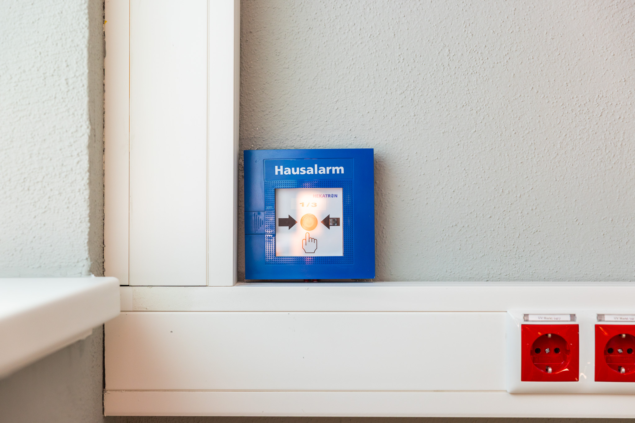 Blauer Hausalarm-Knopf an einer weißen Wand neben zwei roten Steckdosen.