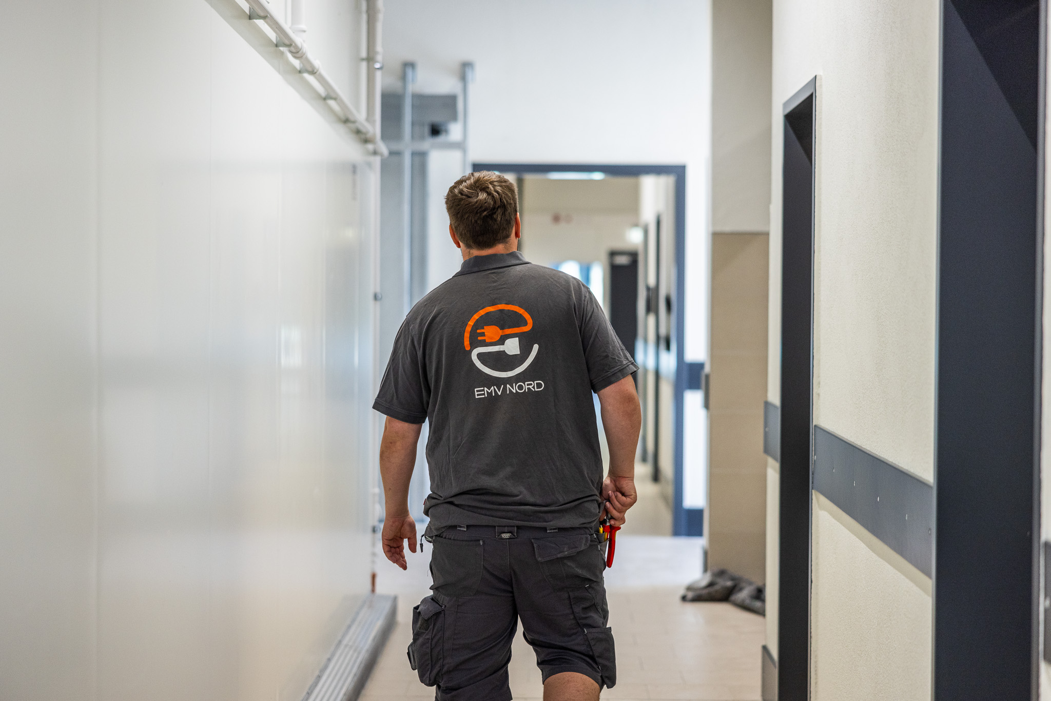Mann in dunkelgrauem EMV NORD T-Shirt geht einen Flur entlang, Werkzeug in der Hand haltend.