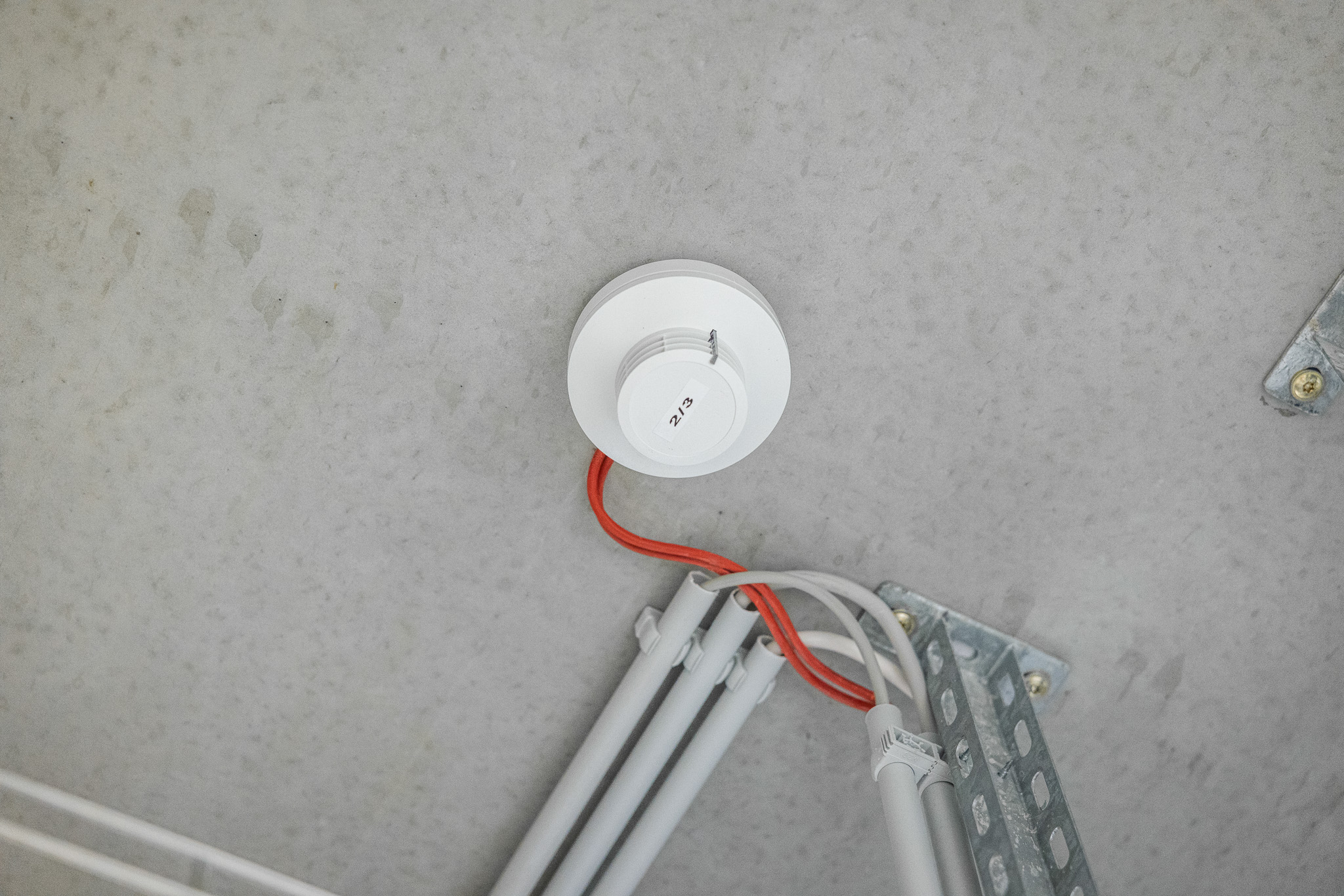Weißer Rauchmelder mit roten und weißen Kabeln an einer grauen Decke montiert.