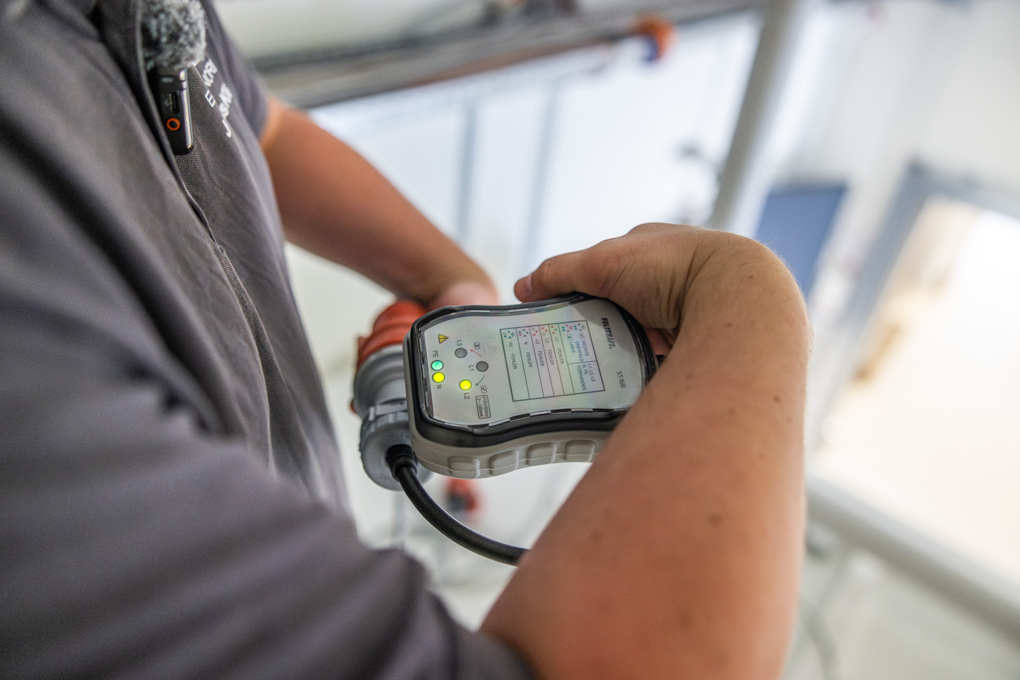 Hand hält Messgerät mit gelben Kontrollleuchten, prüft Kabelverbindung in technischem Umfeld.
