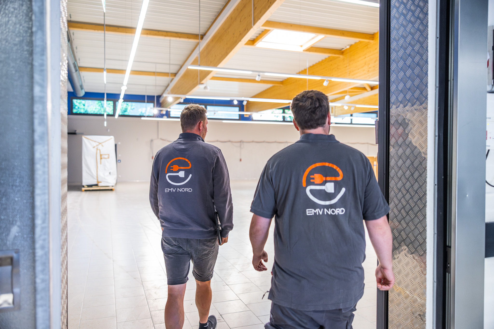 Zwei Männer mit EMV NORD Arbeitskleidung gehen in eine helle, leere Werkhalle.