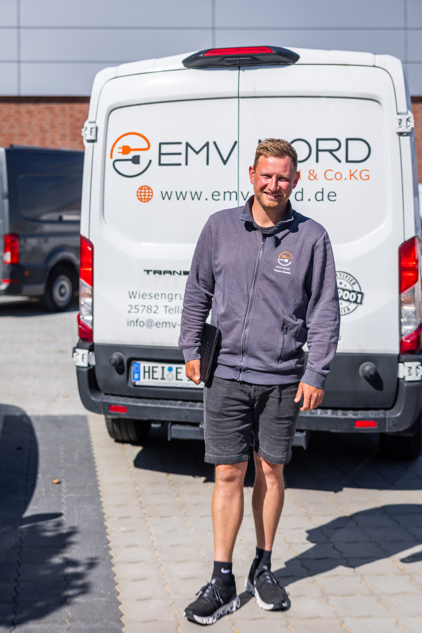 Mann in grauer Jacke und Shorts hält Tablet vor einem weißen Lieferwagen mit Firmenlogo EMV Nord.
