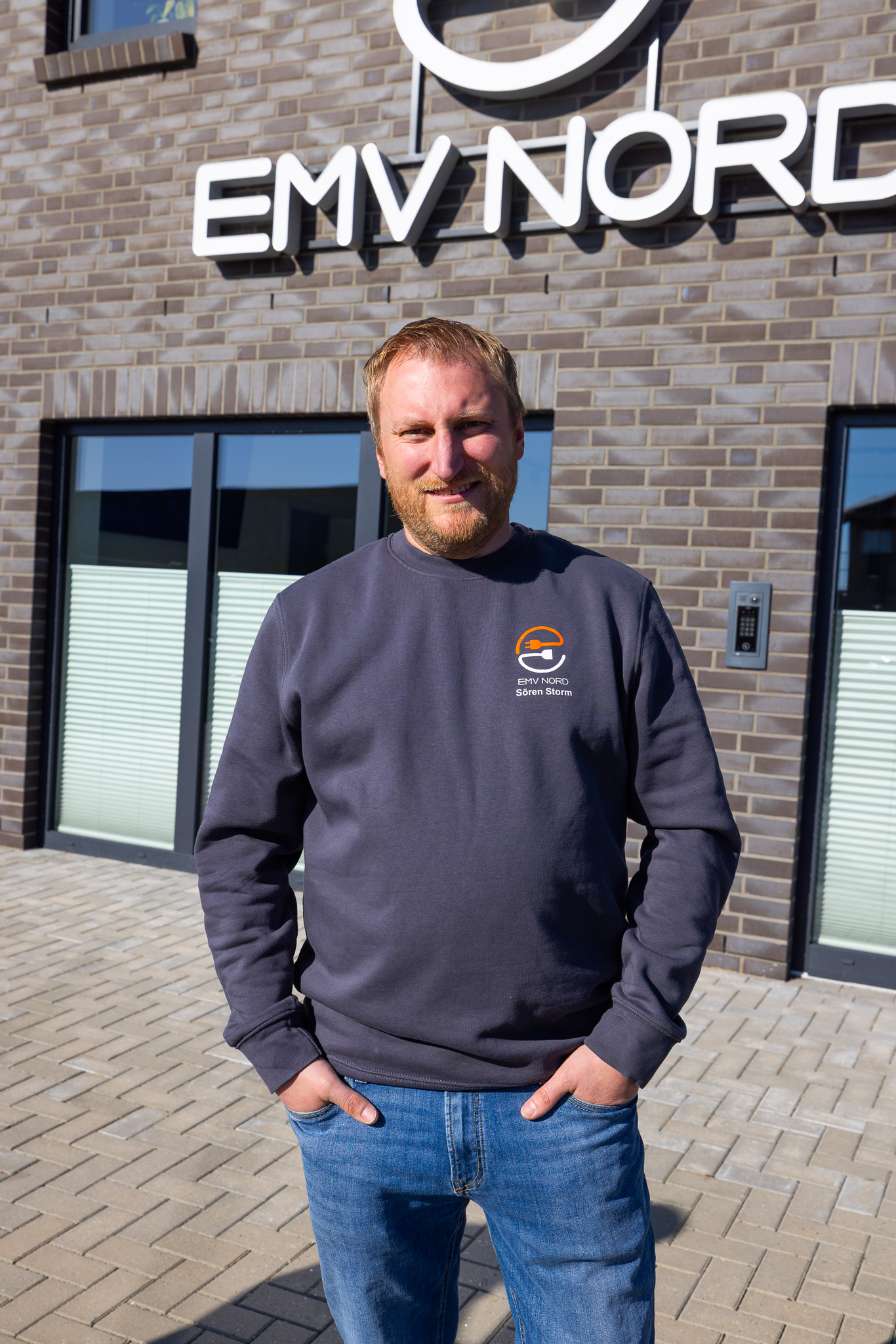 Mann in blauem Pullover und Jeans vor einer Ziegelsteinwand mit EMV NORD Zeichen.
