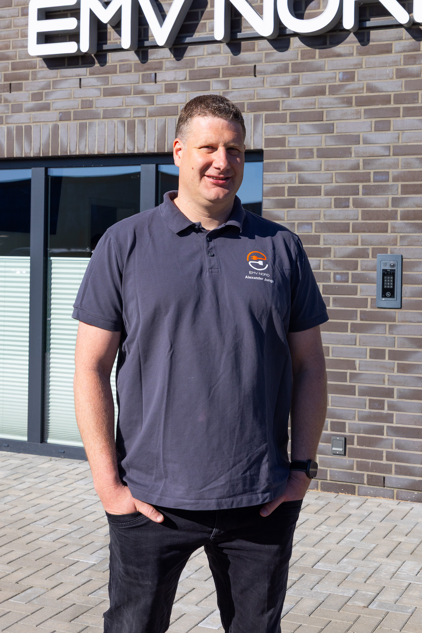 Mann in blauem Poloshirt mit EMV Nord-Logo steht vor Gebäude mit Ziegelmauer.