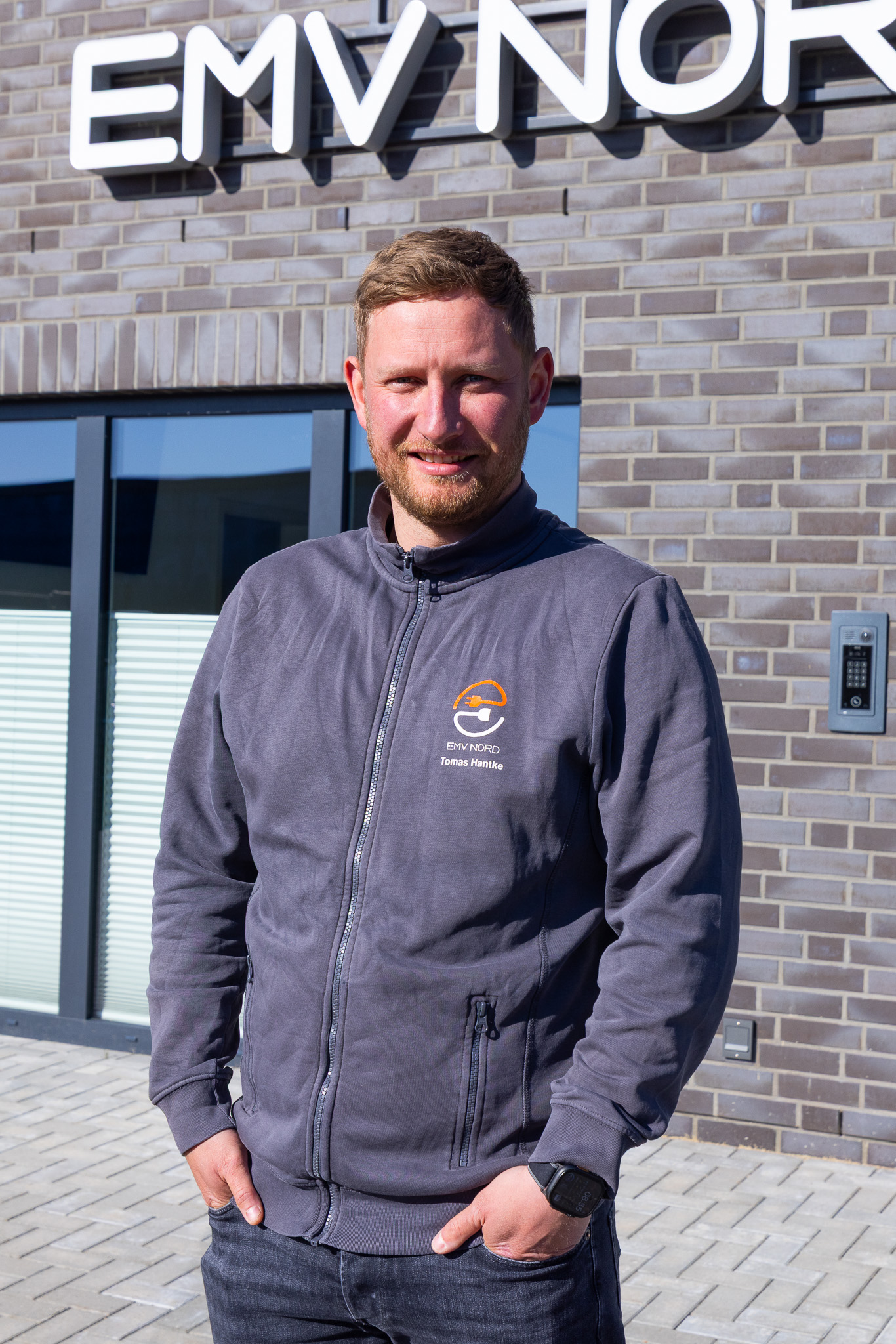 Mann mit grauer Jacke steht vor Gebäude mit EMV NORD Schild und lächelt in die Kamera.