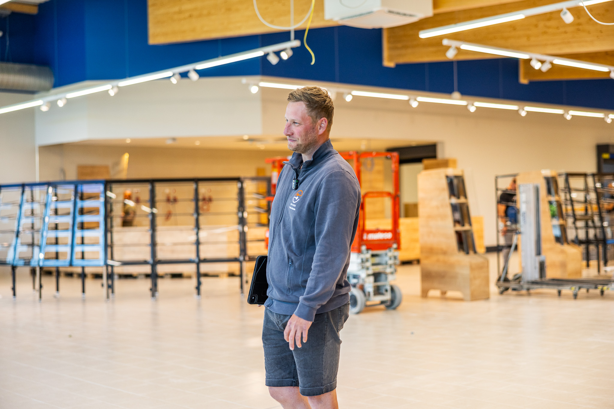 Mann trägt graue Jacke und Shorts in einem hellen, leeren Laden mit Regalen und Holzimitationen.