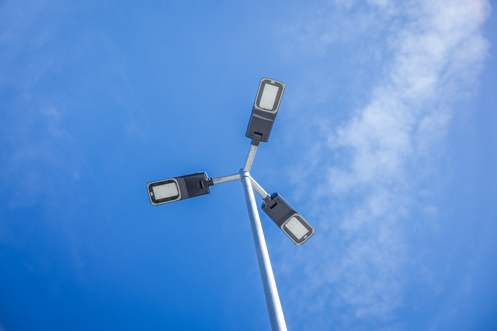 Drei moderne Straßenlaternen auf einem Pfosten vor blauem Himmel mit leichten Wolken.