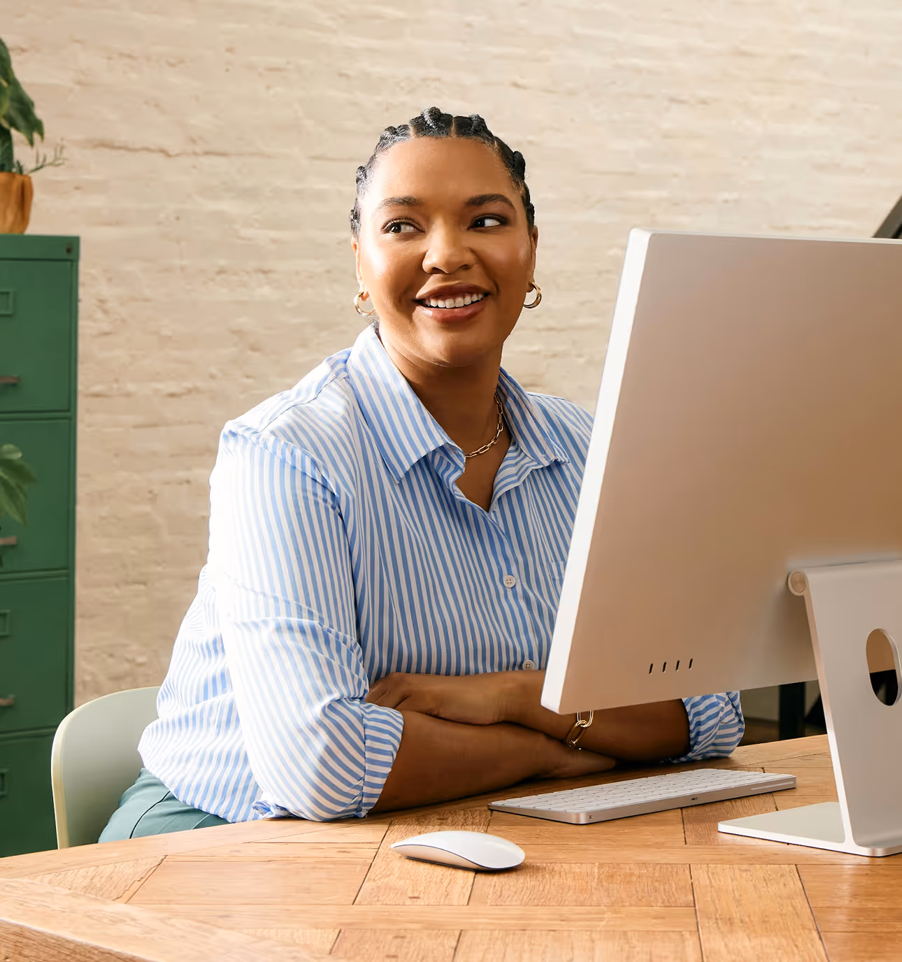 Smiling woman with braided hair sitting at a desk with a computer, looking to the side.