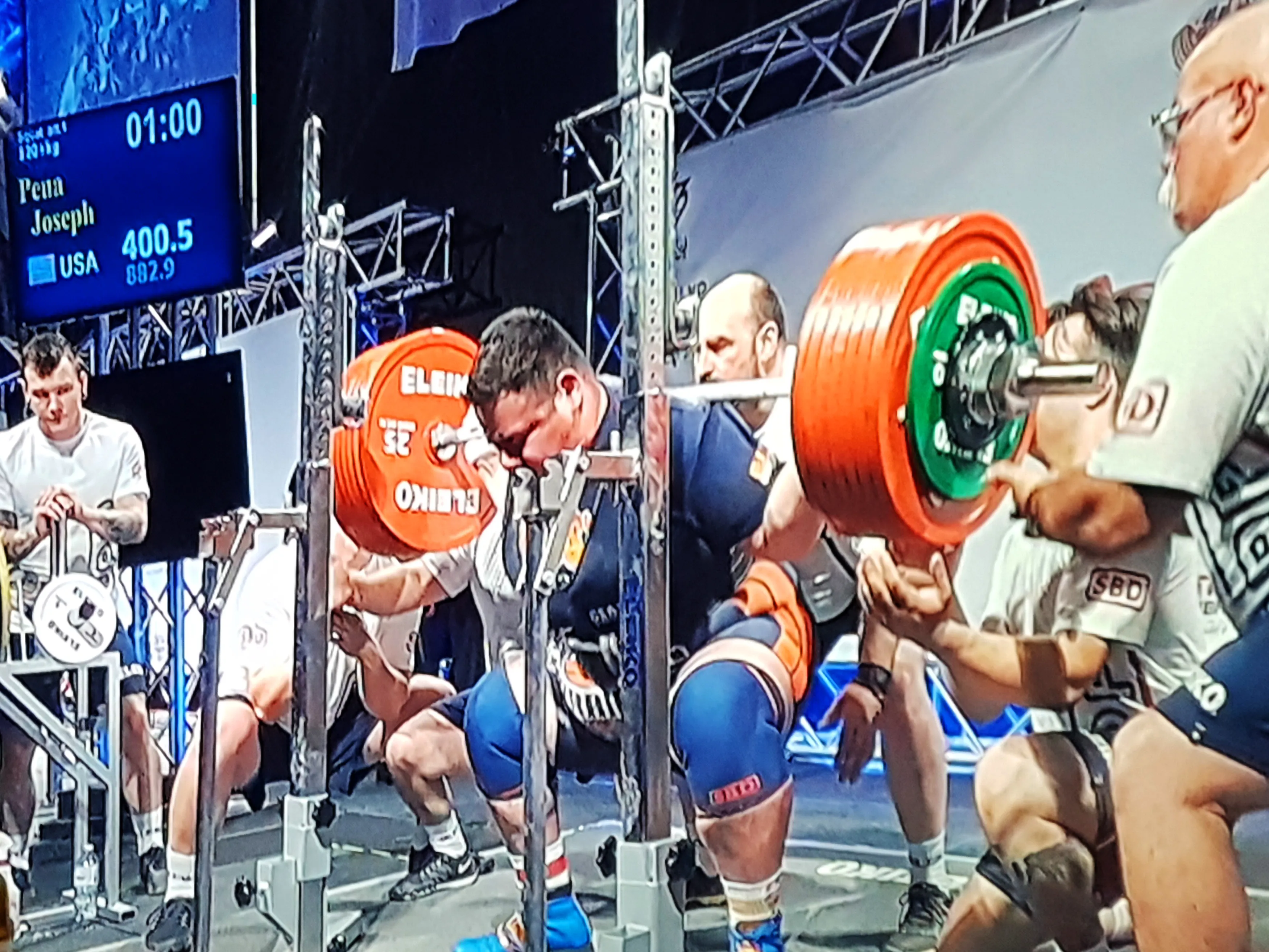 screen showing the powerlifting competition happening. man lifts orange weights