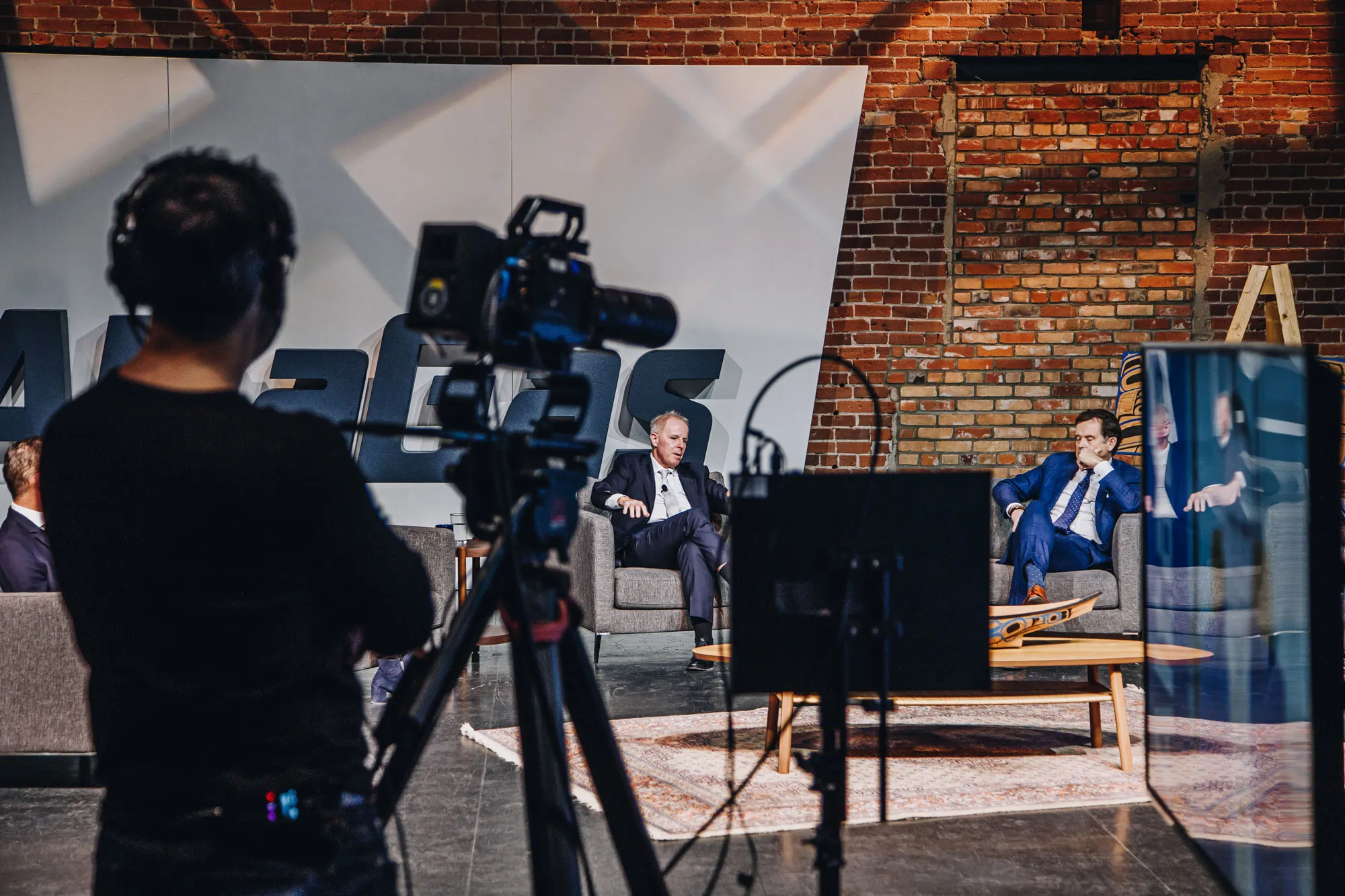Two men in suits sitting on armchairs having a discussion in a recording studio with a cameraman filming them.