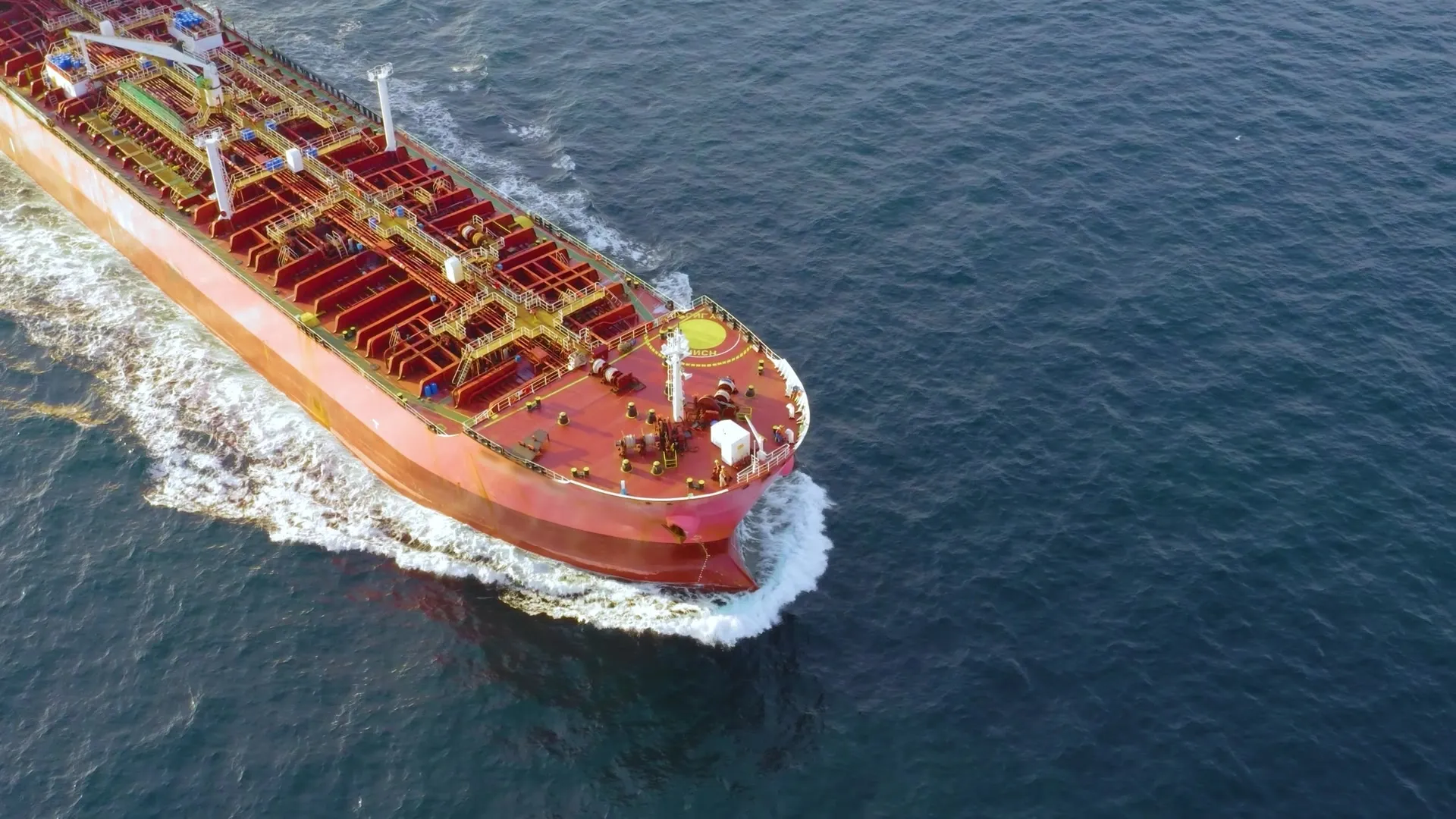 A large red tanker ship sailing through dark blue ocean water, creating white foam at its bow.