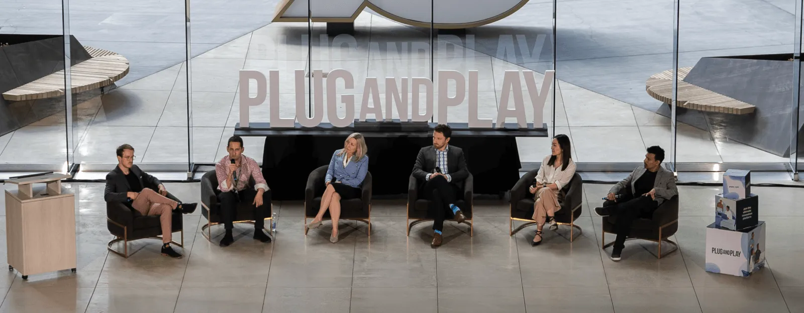 6 speakers sit in arm chairs a semi circle in front of a plug and play sign