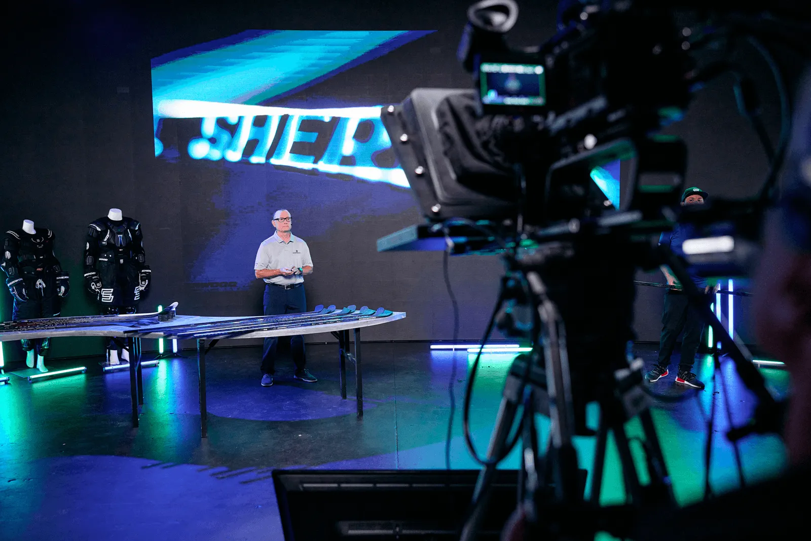 Man standing in studio with hockey sticks on a table and mannequins wearing hockey gear, camera equipment in foreground.