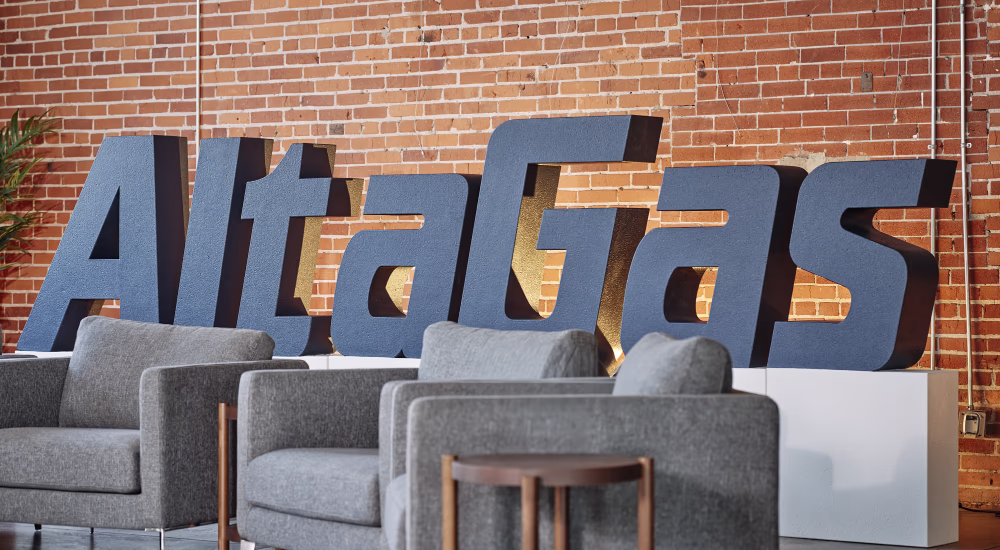 Large textured blue letters spelling 'AltaGas' displayed against a brick wall behind gray upholstered chairs and a small wooden table.