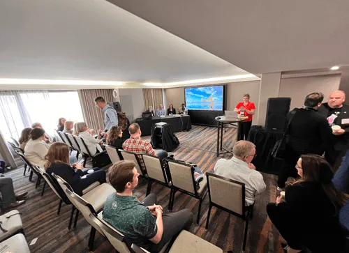 People seated in rows facing a panel of four presenters at the front of a conference room with a screen showing a sailing ship.