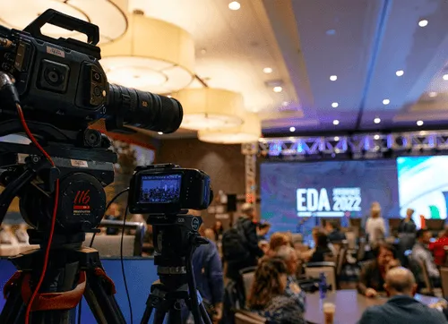 Professional video cameras set up on tripods recording an audience at an indoor event with a screen displaying 'EDA 2022'.
