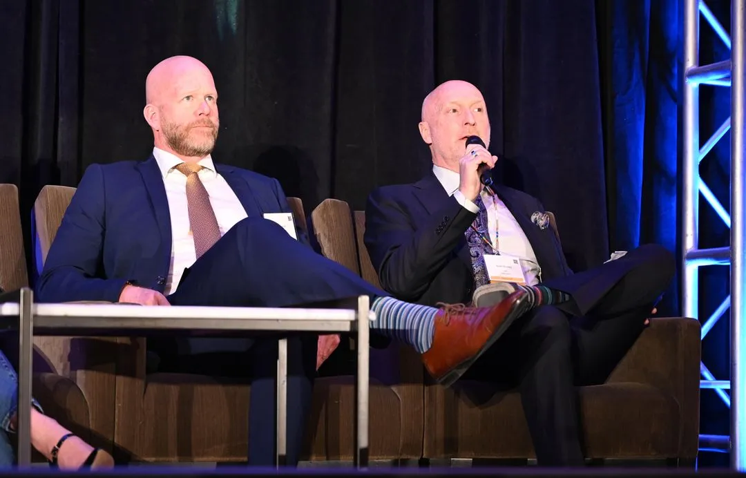 Two men in suits sitting on stage chairs, one speaking into a microphone with his leg crossed showing striped socks and brown shoes.