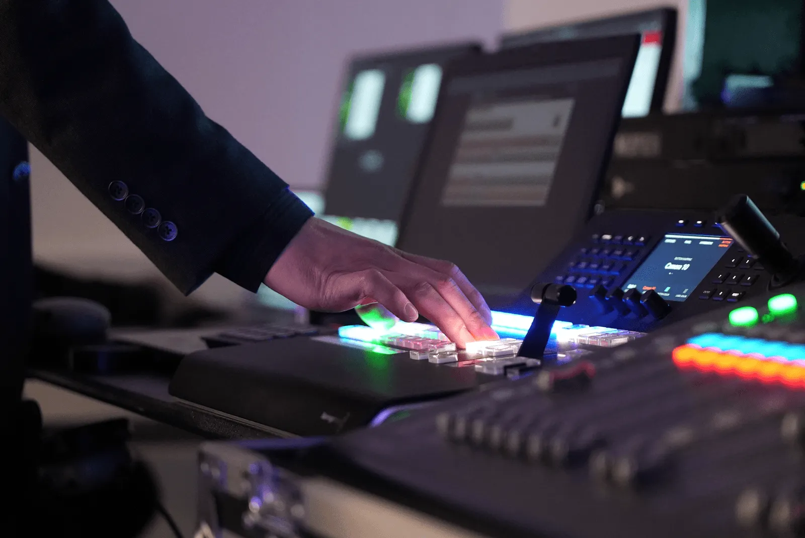 Person's hand operating buttons on a professional video switcher with monitors in the background.