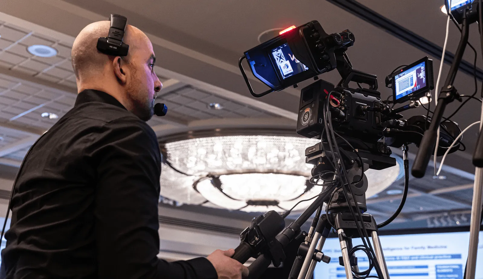 Cameraman wearing headset operating a professional video camera on a tripod indoors with a chandelier in the background.