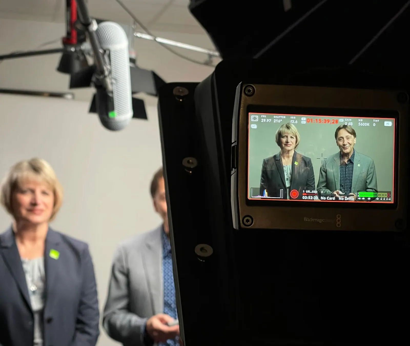 Two people in business attire are being filmed, viewed through a camera monitor with a boom microphone overhead.