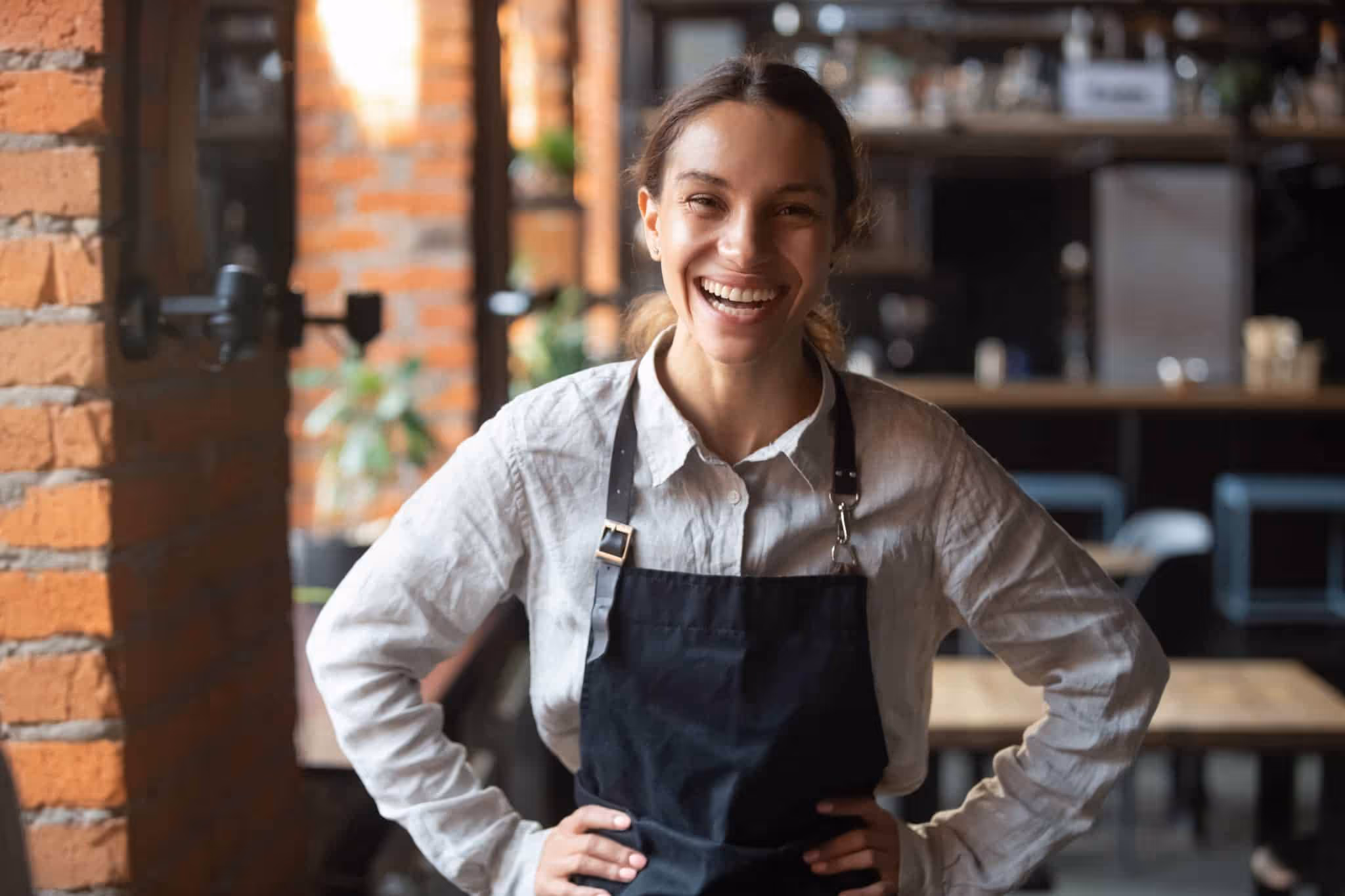Smiling woman wearing a white shirt and black apron standing with hands on hips in a cafe.