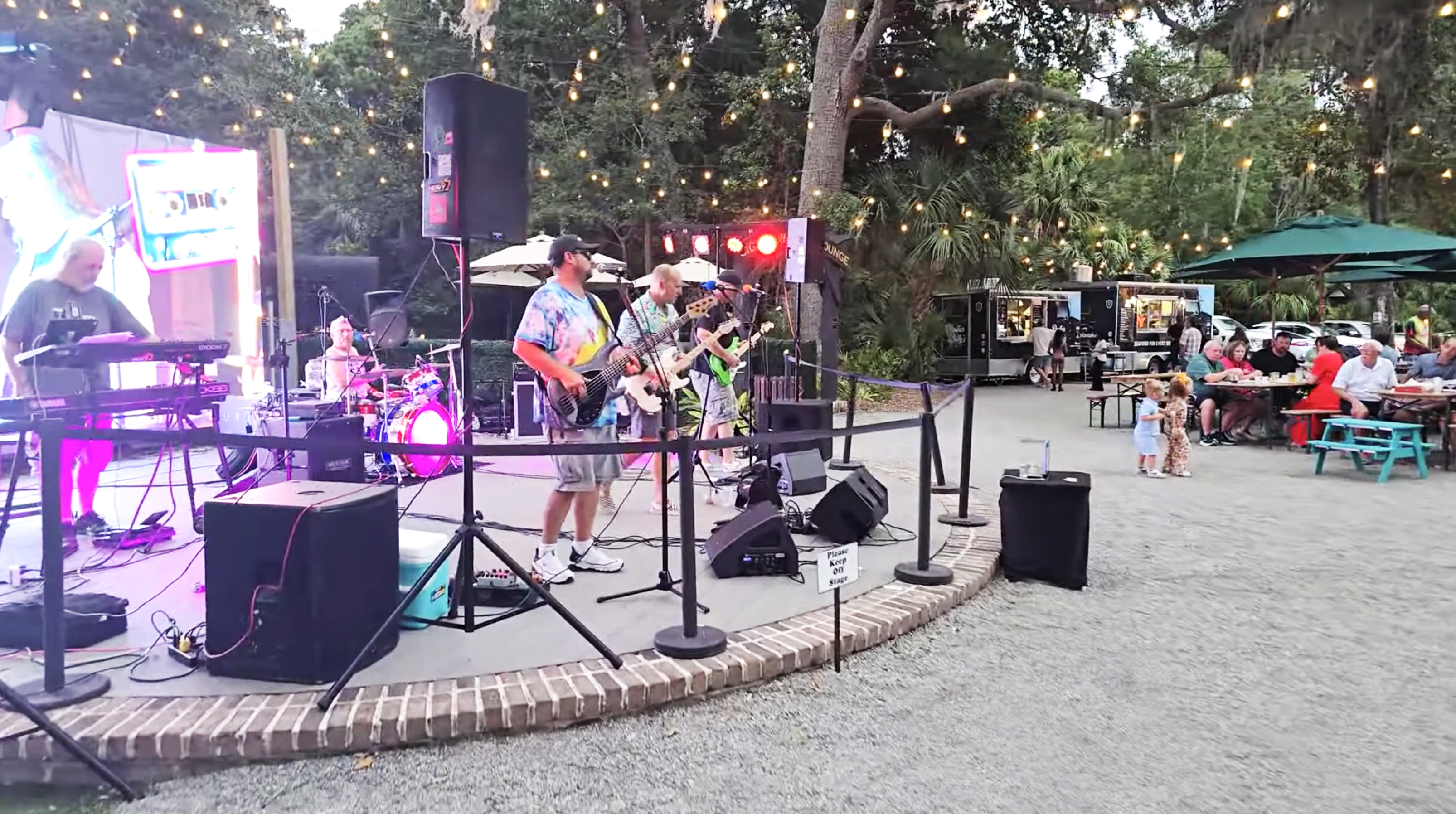 Outdoor band performing on small stage with string lights and seated audience near food trucks.