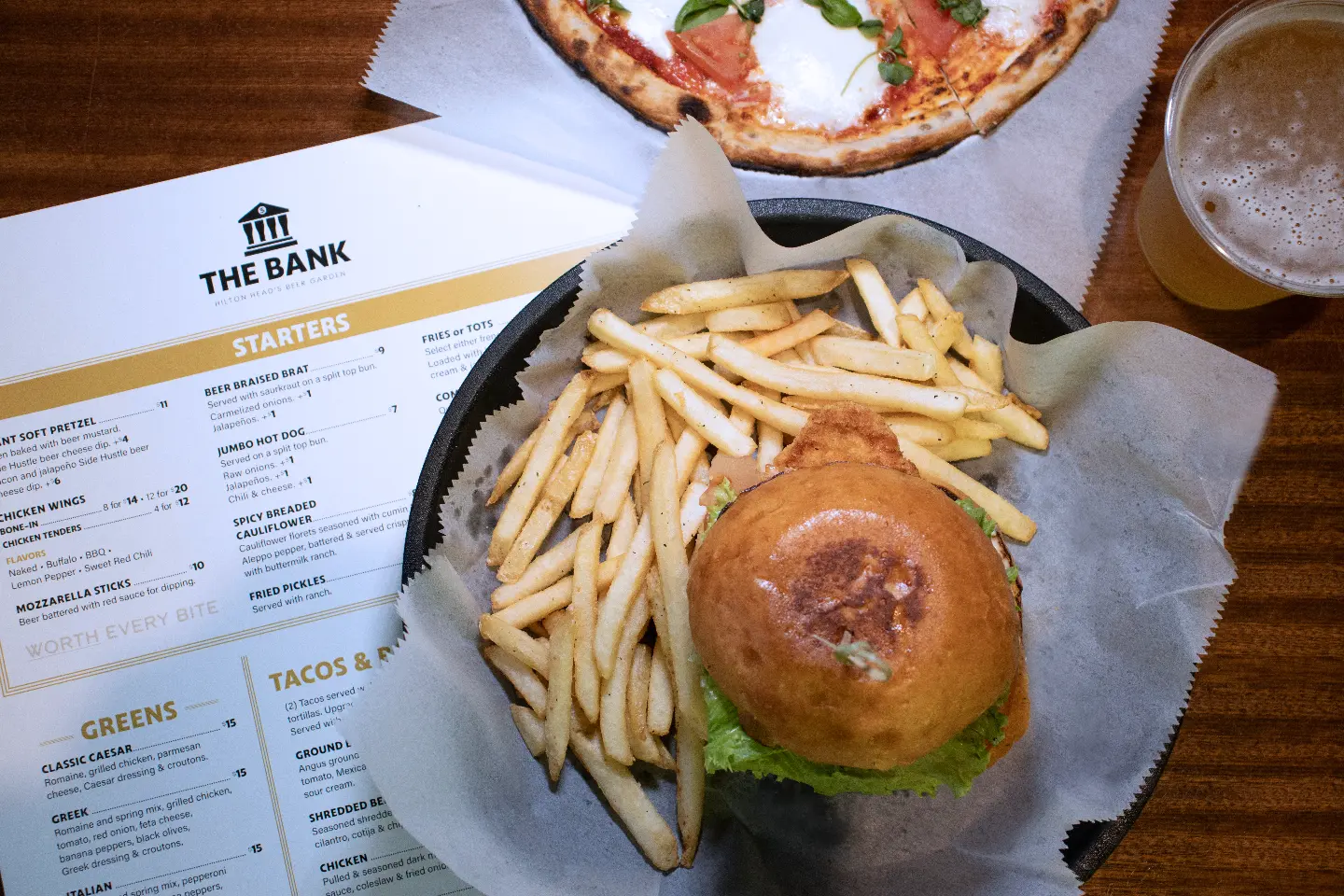 Burger with lettuce and fries on a black tray, pizza slice and beer cup on wooden table.