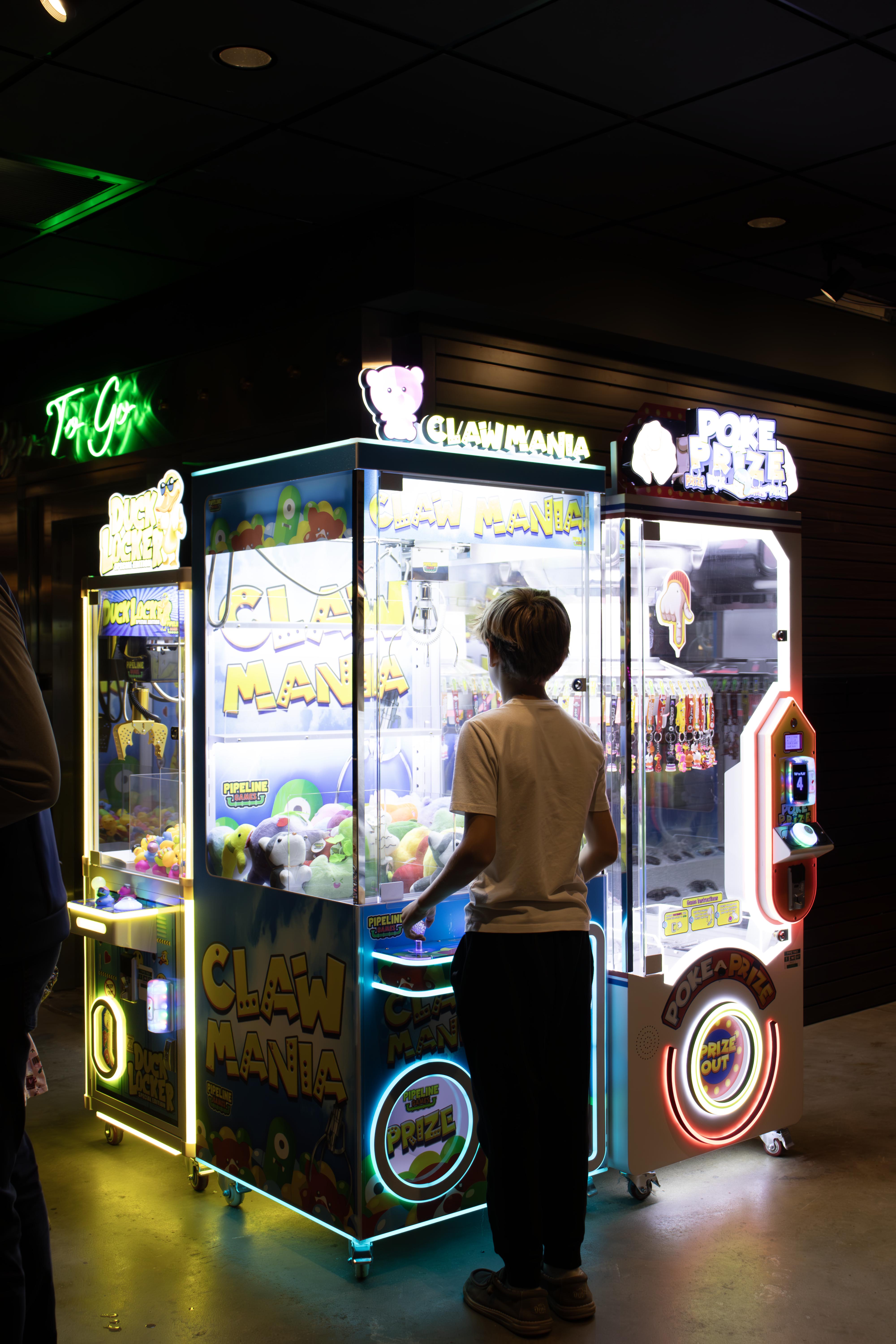 Boy playing a brightly lit arcade claw machine filled with plush toys indoors at night.