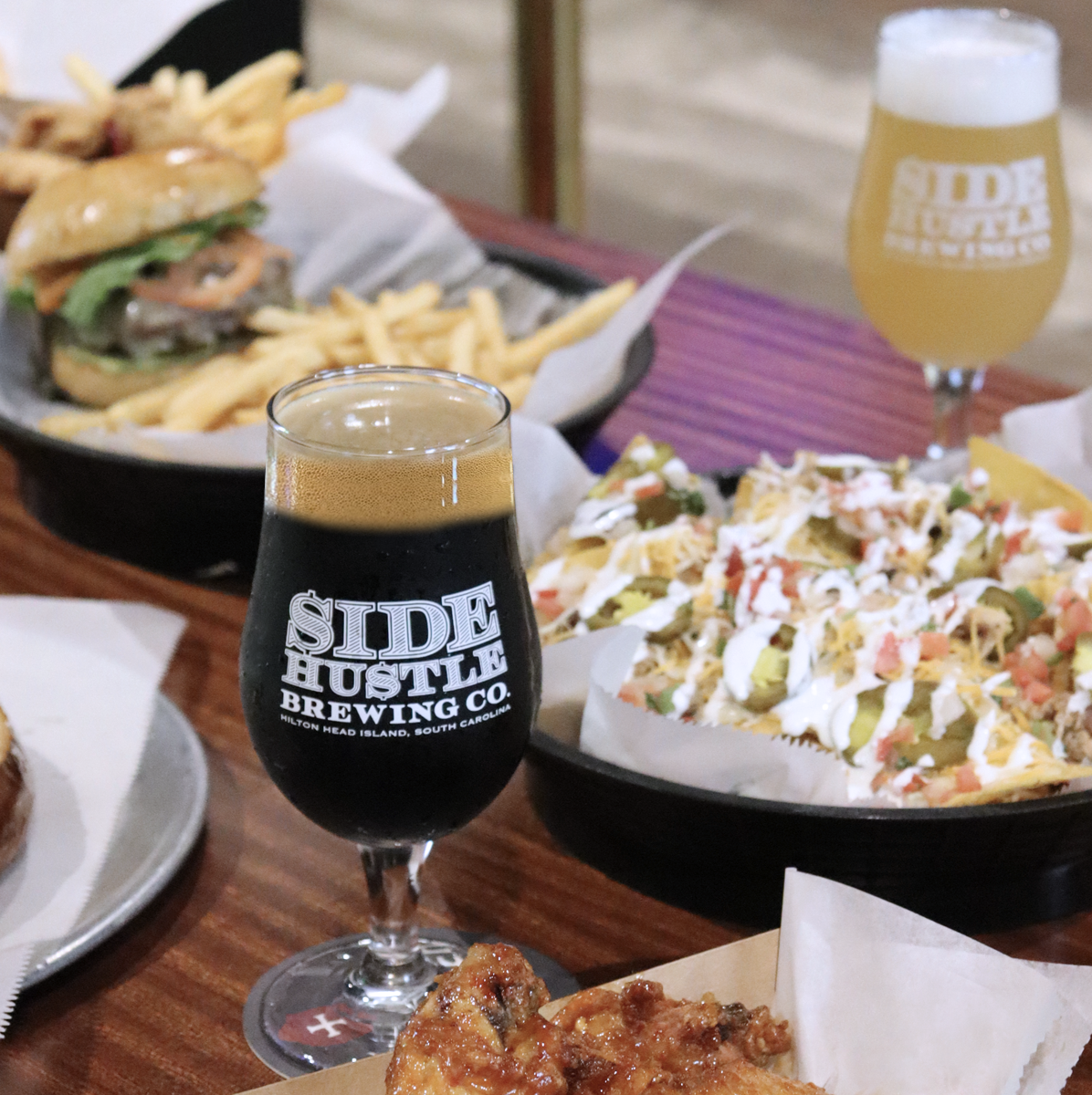 Glass of dark beer from Side Hustle Brewing with wings, nachos, and burger with fries on a wooden table.