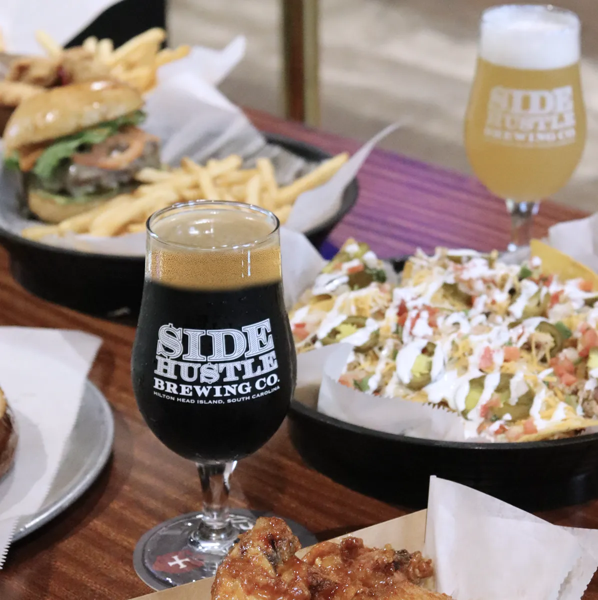 Glass of dark beer from Side Hustle Brewing with wings, nachos, and burger with fries on a wooden table.