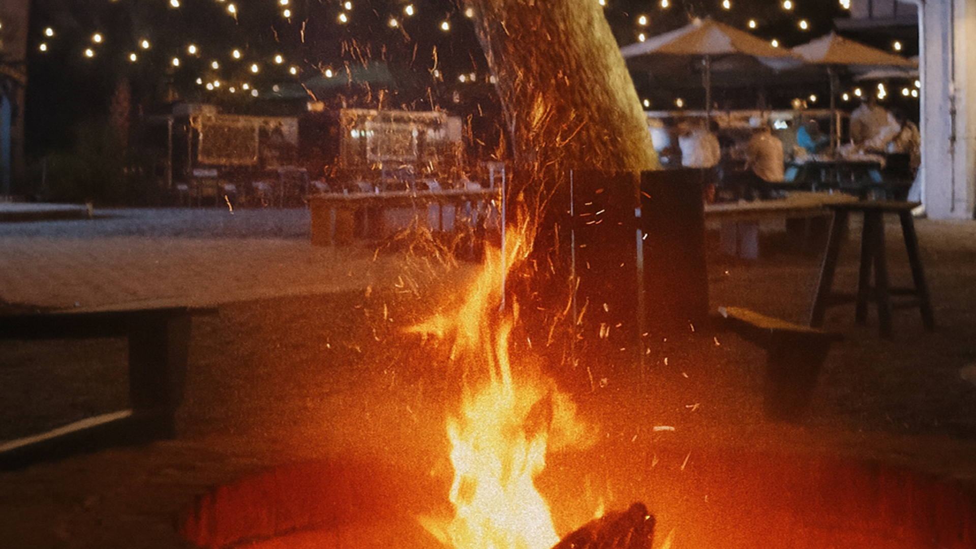 Outdoor firepit blazing with flames and sparks under string lights at night with tables and benches nearby.