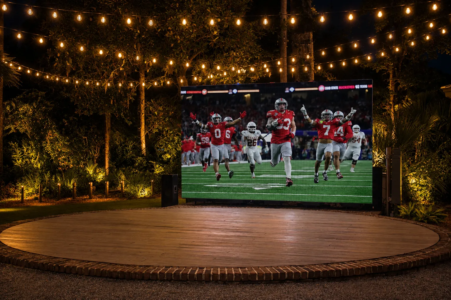 Outdoor nighttime scene with a large screen showing a football game and string lights overhead.