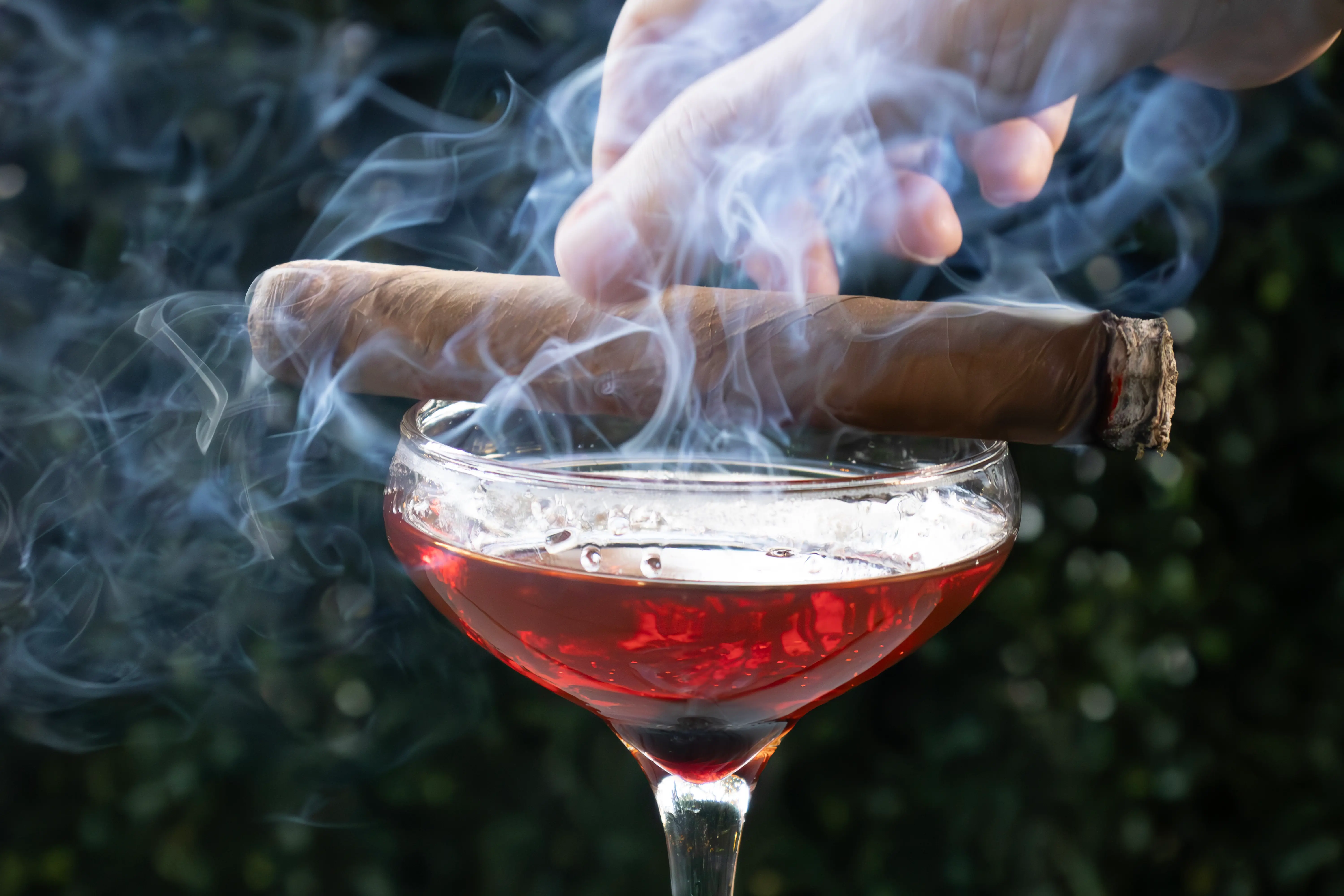 Hand holding a lit cigar above a glass of red cocktail with smoke swirling around.